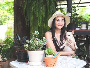 Asian female gardener wearing hat and apron sitting at table with plant pots and gardening tools, holding brown chihuahua dog, smiling and looking at camera.