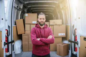 Portrait young happy man courier with folded arms in front of his van with packages ready to be...