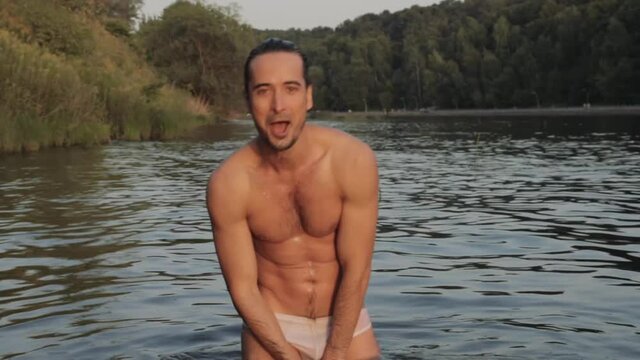 Sexy dripping wet guy fooling around and laughing while bathing in the city river near public park