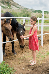 Side view of child girl petting horse at ranch. Kid standing near the white fence in the stable and stroking horse, countryside, Active family weekend lifestyle. Local getaway with kids. Staycation