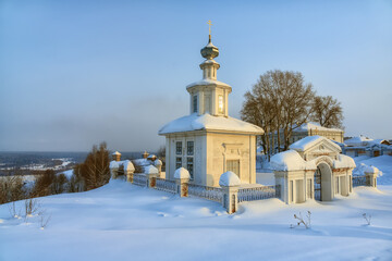 Winter walks in the ancient Ural city of Cherdyn (Ural, Russia). A church, old houses and branchy linden trees rise above a deep snow-covered field. The river valley is visible under the mountain 