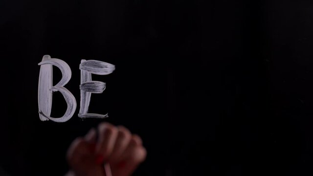 Hand is writing message. The phrase 'Be free' written on glass using brush and white paint. Freedom concept. Black background. 4k footage