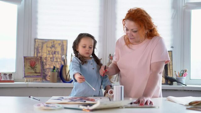 Mother helps hes little daughter to do her drawing or drawing together concept. Happy creative family. Caring caucasian mother or babysitter drawing with colored watercolor paints teaching child girl