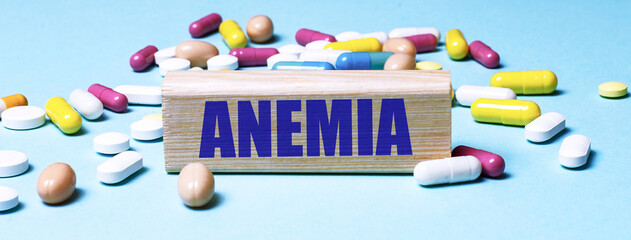 A wooden block with the word ANEMIA stands on a blue background among multi-colored pills. Medical...