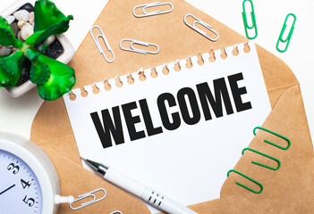 On a light background, an open envelope, a white alarm clock, a green plant, white and green paper clips, a white pen and a sheet of paper with the text WELCOME