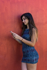 Young woman chatting with her smartphone. It is leaning against a maroon wall. She wears a short dress. It is located in Las Americas, Tenerife