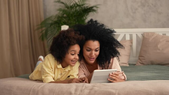 African American woman and litle girl talking on video call using digital tablet. Mother and daughter posing, lying on the bed in a bedroom with a bright interior. Slow motion ready, 4K at 59.97 fps.