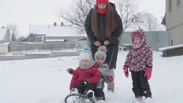 vacation, game, family concept - slo-mo authentic three happy preschool toddler kids siblings with young wonan mom sledding and play with snow. snowfall in cold season weather in winter yard outdoors