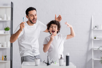 Cheerful arabian man with toothbrush near singing son in bathroom