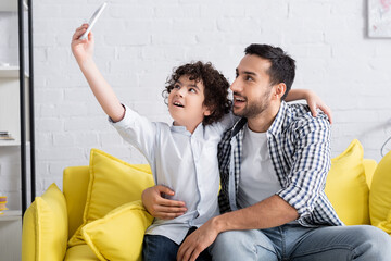 Obraz premium happy muslim father and son taking selfie on sofa at home