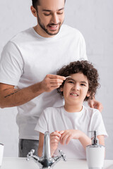 Fototapeta premium Smiling arabian man holding tweezers near eyebrow of confused son in bathroom