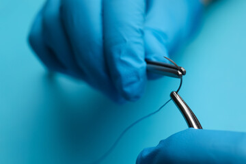 Professional surgeon holding forceps with suture thread on light blue background, closeup. Medical...