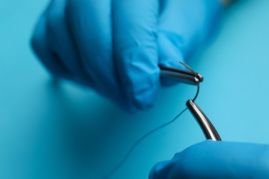 Professional surgeon holding forceps with suture thread on light blue background, closeup. Medical equipment