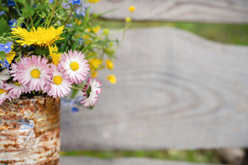a bouquet of wildflowers of forget-me-nots, daisies and yellow dandelions in full bloom in a rusty rustic jar against a background of wooden planks in nature. cottagecore scene. space for text