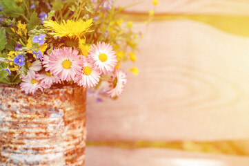 a bouquet of wildflowers of forget-me-nots, daisies and yellow dandelions in full bloom in a rusty rustic jar against a background of wooden planks in nature. cottagecore scene. space for text. flare
