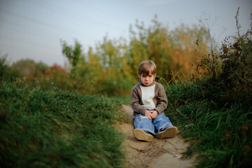 Naklejka na ściany i meble Sad boy child sits on a path in the park in the forest, a lonely child. Concept Children's emotions and psychology