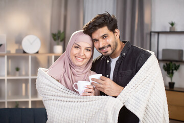 Romantic relationship. Cute Arabian Muslim couple standing together under warm blanket in the living room and drinking coffee together.