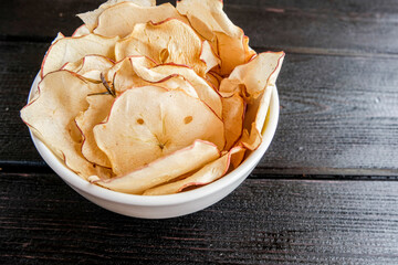 Chips of fresh apples with cinnamon are on a white plate on a background of black wooden boards. Organic apple chips. Dried fruits. Healthy sweet snack. Dehydrated and raw food. Copy space.