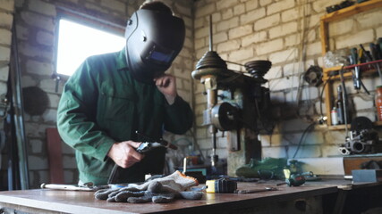 Young mechanic in protective mask holding broken welding machine and trying to solder some metal details at garage. Male welder working at his workshop. Concept of maintenance service. Close up