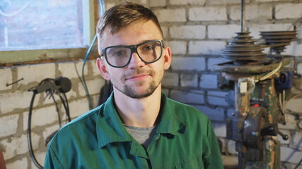 Handsome young repairman in protective glasses looking into camera at garage. Portrait of happy professional mechanic in uniform at workshop. Concept of maintenance service. Close up Dolly shot