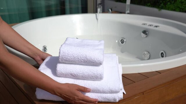 Woman takes away stack of four folded clean soft white towels laying near empty whirlpool bath