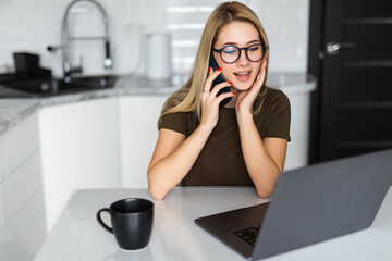 Young woman with laptop using cellular phone in the kitchen