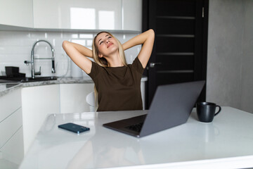 Young woman relaxing in kitchen leaning back in a chair with her hands clasped behind her neck and her eyes closed in front of a laptop