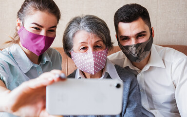 Adult children visit an elderly mother and take a selfie at home on the couch-A woman with gray hair is photographed with young people wearing masks on her face in quarantine - Caring for an old woman - Powered by Adobe