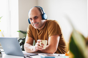 A man at home with headphones and a cup of tea looking at a laptop with pleasure and listening to...