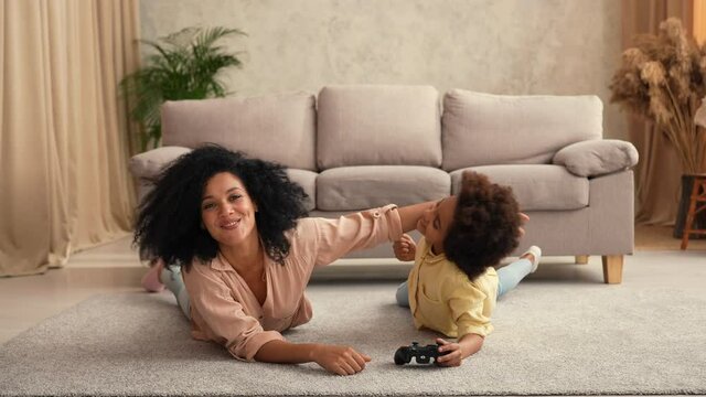 Little African American girl plays game on wireless joystick and rejoices victory. Mother and daughter lying on floor in room with light home interior. Slow motion ready, 4K at 59.97 fps.