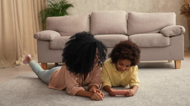 Little African American girl plays game on smartphone and rejoices victory. Mother and daughter posing, lying on floor in room with light home interior. Slow motion ready, 4K at 59.97 fps.