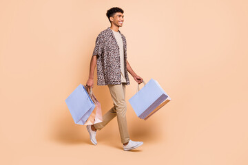 Full body profile portrait of cheerful dark skin guy look empty space carry bags isolated on beige...