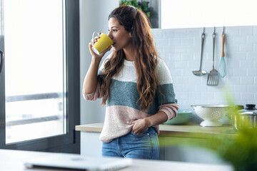 Beautiful young woman drinking a cup of coffee while looking forward standing next to the window in the kitchen at home.