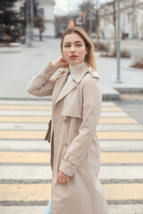 Young beautiful blonde hair woman in fashion clothes: beige trench coat, black boots and jeans crossing the road street. City lifestyle portrait.