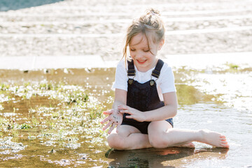 Cute cheerful girl playing in fountain. Kid in denim overall having fun in summer park