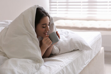 Beautiful young woman covered with warm blanket enjoying hot drink on bed at home. Space for text