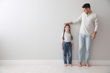 Father measuring little girl's height near light grey wall indoors. Space for text