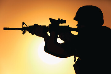 Silhouette of modern army infantry soldier standing on background of setting sun and aiming assault...