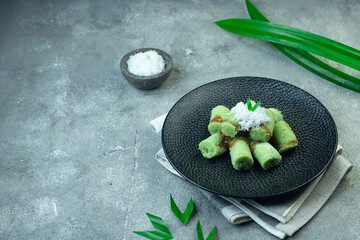 Kue Putu or Indonesian Putu bamboo Cake, traditional cake made from rice flour, palm sugar, and grated coconut. Served with black ceramic plate, garnished with pandanus leaves on grey  background.