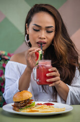 Asian young woman drinking red watermelon smoothie and eating unhealthy burger and french fries food in cafe or restaurant