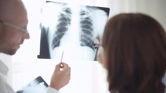 4K Two doctors examining X-ray scans of person's chest against bright light. Male doctor pointing at scan with pen. Medical concept.