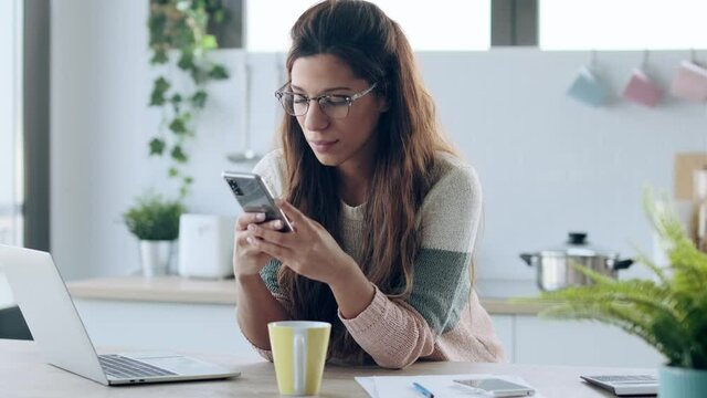 Video of concentrated young business woman using her mobile phone while working with laptop in the kitchen at home.