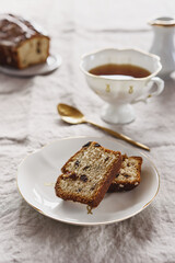 Fototapeta na wymiar Slice of Pound Loaf Cake with raisins on white porcelain plate and cup of tea on greige linen tablecloth. Selective focus