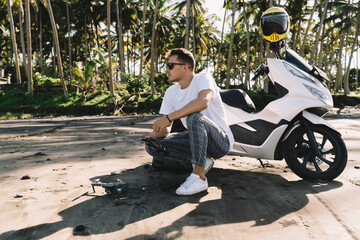 Dreamy young man squatting by motorcycle with drone controller on palms background