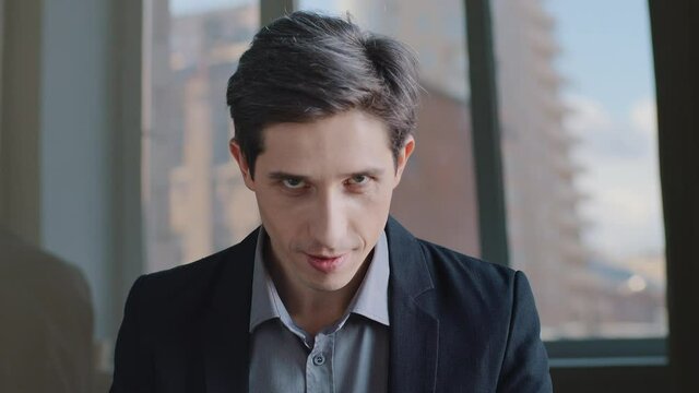 Portrait of Caucasian millennial business man sitting in modern office contemplating insidious plan, looking with cunning at camera. One 30s guy professional successful boss bad human going to do evil