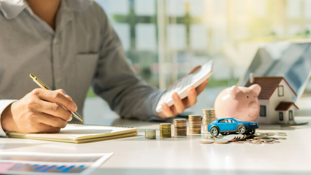 Hand holding a pen with stacked coins and a wooden house model with a car or real estate brokerage idea. Insurance or real estate loans Business house investment ideas