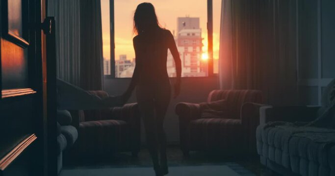 Young woman takes off her summer dress and dances near the wide window in the apartment at sunset