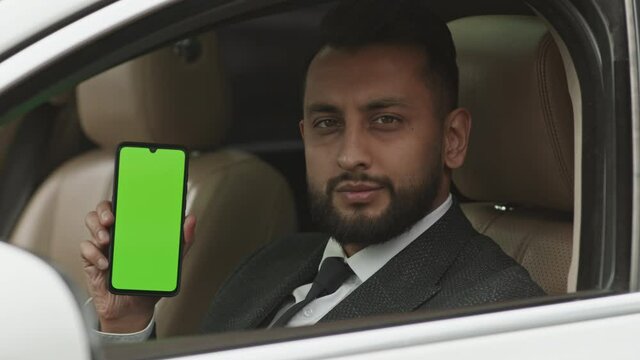 Shoulder-up POV Of Young Mixed-Race Man Wearing Formal Clothing, Sitting On Drivers Seat, Showing Green-screened Smartphone And Looking Outside Car On Camera