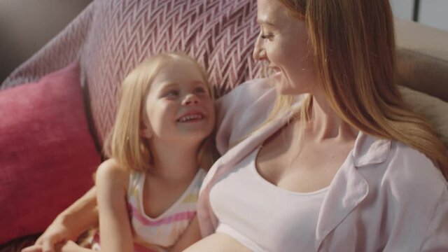 High angle shot of loving pregnant mother and cute little daughter bonding together on sofa at home, looking at each other and smiling