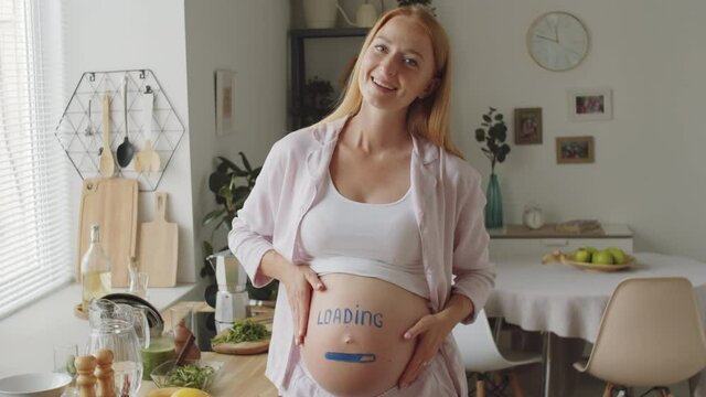 Medium shot of cheerful expectant mother standing at home, looking at camera, smiling and stroking her pregnant belly with loading sign and painted progress bar